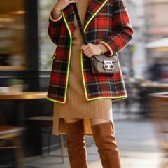 A realistic, high-quality fashion editorial photo of a stylish female model standing in a modern urban setting with soft, natural daylight. The model wears a camel knit midi dress that falls to mid-calf, layered under a bold, statement-length coat with a striking plaid pattern, hitting at the knee to elongate the frame. The coat features neon trim accents for a pop of color. She pairs the outfit with over-the-knee boots in a sleek suede or leather finish and a small crossbody bag with metallic hardware to add a touch of shine. Keep jewelry minimal: a single delicate ring on one hand. The pose should convey confidence and elegance—one hand resting lightly on the coat lapel, the other holding the crossbody strap. Background should be softly blurred but hints of a chic city street or a stylish cafe to emphasize a fashion editor vibe. No visible text on the image.