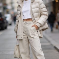 A fashion-forward, high-resolution editorial portrait of a confident woman modeling sporty-chic layering in a city street setting on a mild day. She stands relaxed with one hand in a pocket, looking slightly off-camera. Outfit: a lightweight puffer jacket over a white cropped tee, layered with a soft neutral cardigan, and wide-leg trousers in a complementary tone. White sneakers with a subtle elevated sole complete the look, paired with a slightly cropped jacket that reveals the longer top underneath to create vertical layering. The proportions are balanced: cropped jacket over a longer top, cardigan peeking from beneath the puffer for warmth-to-cool transition. Accessories are minimal: a simple neutral tote and small hoop earrings. Lighting is natural with soft diffused daylight and a shallow depth of field to gently blur the urban background, highlighting texture on fabrics (puffer, knit, denim/trouser) and the model’s confident, approachable expression. The overall mood is effortless, wearable, and versatile for unpredictable weather and busy days. No text in the image.